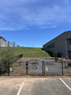 A black wrought iron fence and gate enclose a paved lot against a grassy hillside.